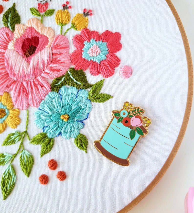May include: A white embroidery hoop with a floral design embroidered in pink, yellow, green, and blue thread. A gold-tone enamel pin of a spool of thread with flowers on top is attached to the hoop.