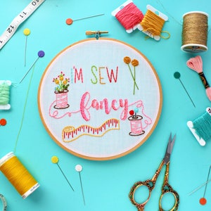May include: A white embroidery hoop with the text "I'M SEW fancy" embroidered in pink, green, and yellow thread. The hoop is surrounded by various sewing supplies, including spools of thread, needles, and scissors.