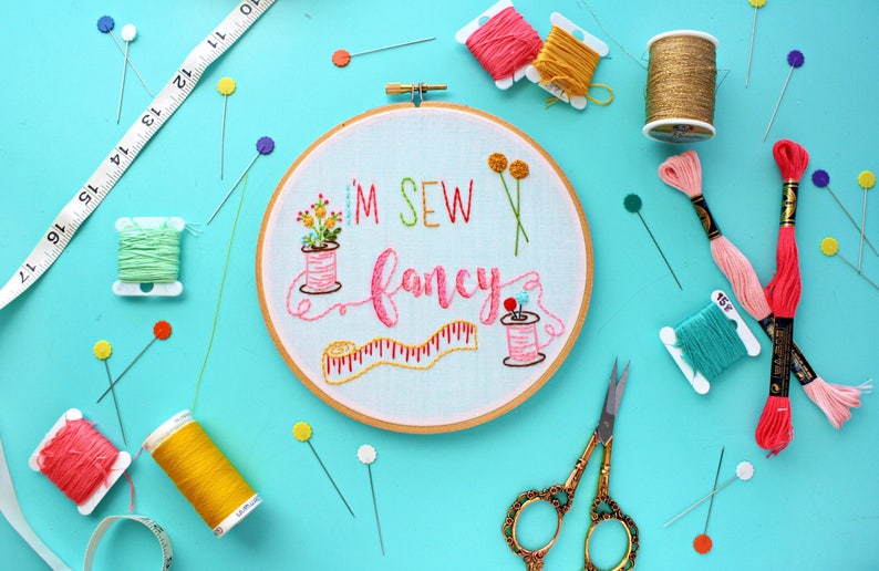 May include: A white embroidery hoop with the text "I'M SEW fancy" embroidered in pink, green, and yellow thread. The hoop is surrounded by various sewing supplies, including spools of thread, needles, and scissors.