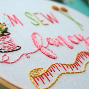 May include: Embroidered hoop art featuring the text "I'm Sew Fancy" in pink thread. The hoop also includes a spool of thread, a measuring tape, and a small bunch of flowers.