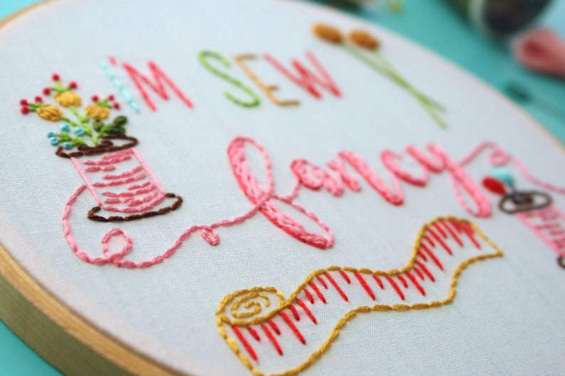 May include: Embroidered hoop art featuring the text "I'm Sew Fancy" in pink thread. The hoop also includes a spool of thread, a measuring tape, and a small bunch of flowers.