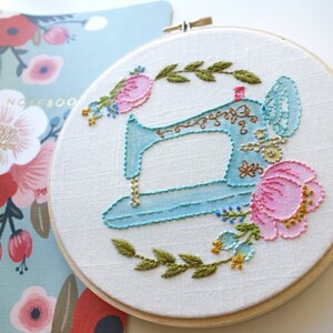 May include: A blue sewing machine embroidered on a white fabric with pink and green flowers. The embroidery is in a hoop.