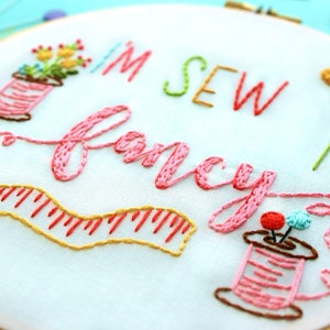 May include: Embroidered hoop art featuring the text "I'm Sew Fancy" in pink thread. The design includes two spools of thread, a measuring tape, and a small flower arrangement.