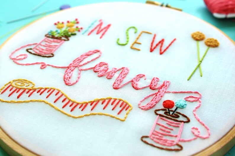 May include: Embroidered hoop art featuring the text "I'm Sew Fancy" in pink thread. The design includes two spools of thread, a measuring tape, and a small flower arrangement.