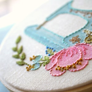 May include: A white embroidery hoop with a blue and white embroidered shoe, a pink flower, and green leaves.