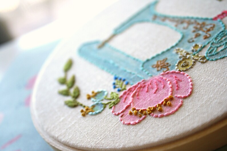 May include: A white embroidery hoop with a blue and white embroidered shoe, a pink flower, and green leaves.