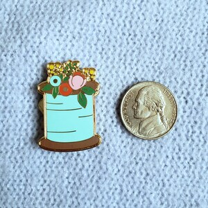 May include: A gold-tone enamel pin featuring a spool of thread with a blue body and a brown base. The spool is topped with a cluster of colorful flowers, including pink, red, yellow, and blue.