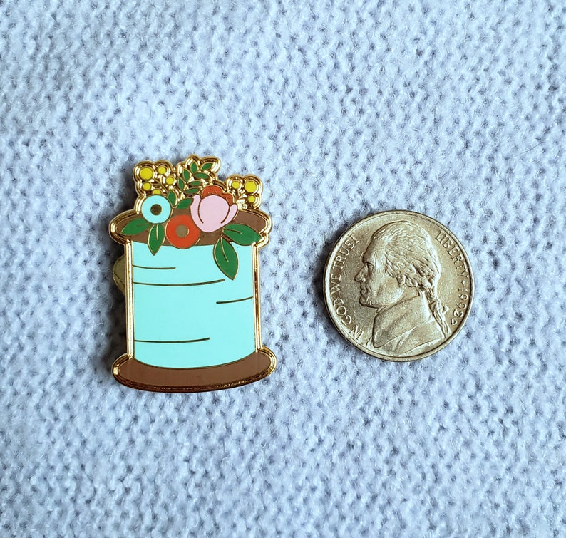 May include: A gold-tone enamel pin featuring a spool of thread with a blue body and a brown base. The spool is topped with a cluster of colorful flowers, including pink, red, yellow, and blue.