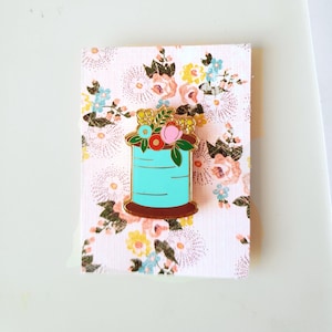 May include: A gold-tone enamel pin featuring a spool of turquoise thread with a floral arrangement on top. The flowers are pink, red, yellow, and blue.