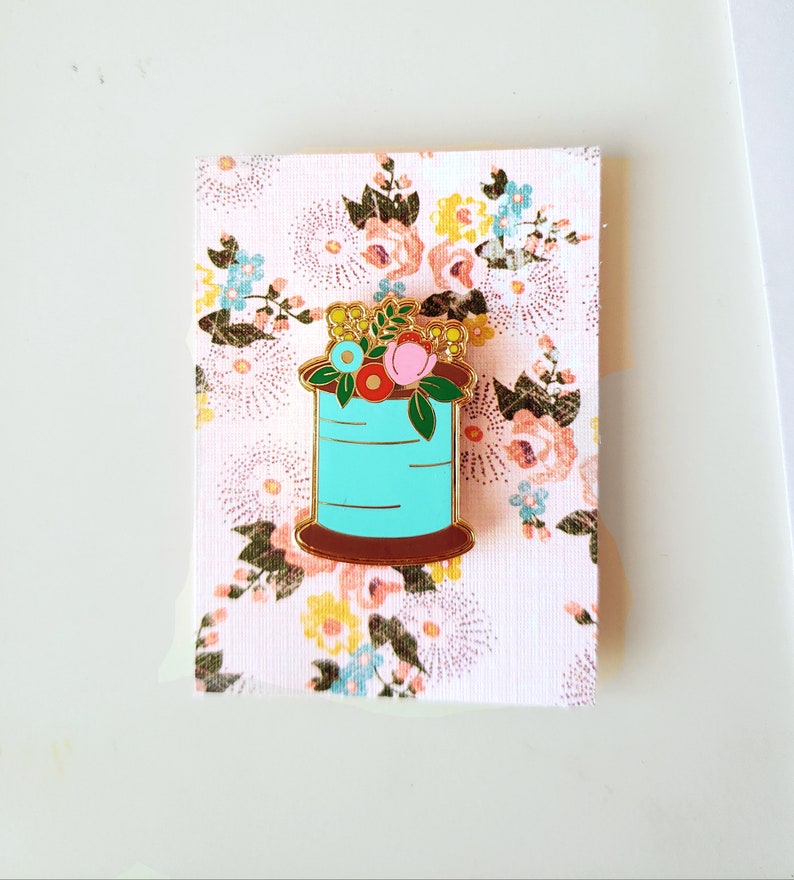 May include: A gold-tone enamel pin featuring a spool of turquoise thread with a floral arrangement on top. The flowers are pink, red, yellow, and blue.
