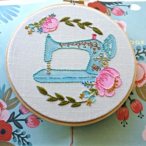 May include: A blue sewing machine embroidered on white fabric in a wooden embroidery hoop. The machine is surrounded by pink and green flowers.