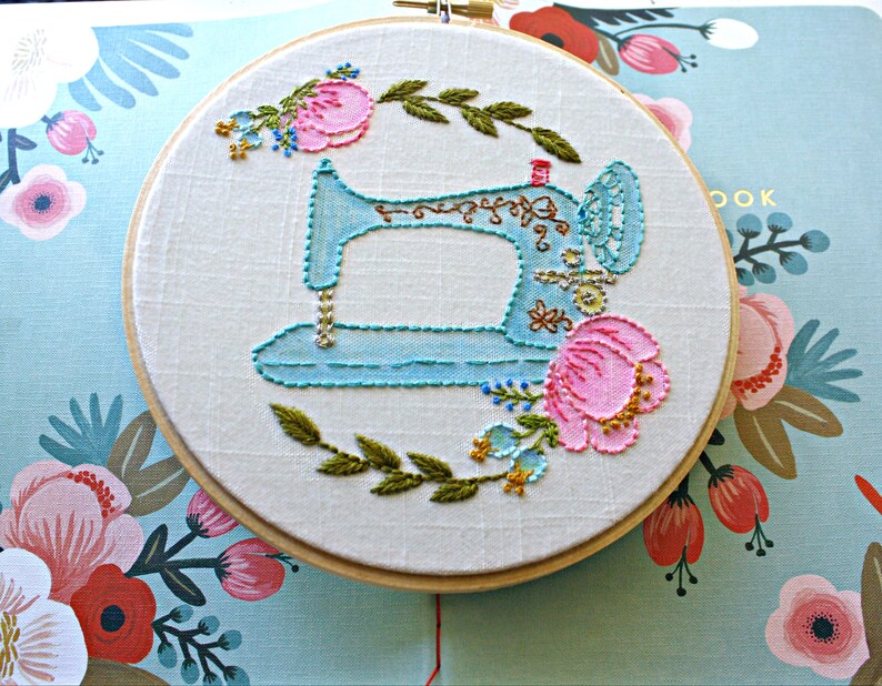 May include: A blue sewing machine embroidered on white fabric in a wooden embroidery hoop. The machine is surrounded by pink and green flowers.