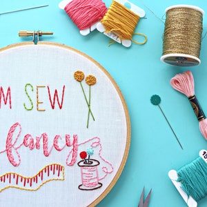 May include: Embroidered hoop art with the text "I'm Sew Fancy" in colourful thread. The hoop features a spool of thread, a measuring tape, and flowers.