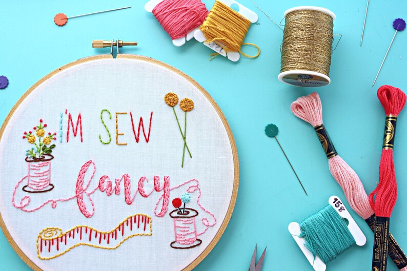 May include: Embroidered hoop art with the text "I'm Sew Fancy" in colourful thread. The hoop features a spool of thread, a measuring tape, and flowers.