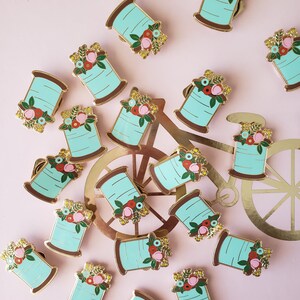 May include: A collection of 18 gold-tone enamel pins featuring a spool of thread with a floral design. The spools are turquoise blue with a gold-tone rim and a variety of colorful flowers.
