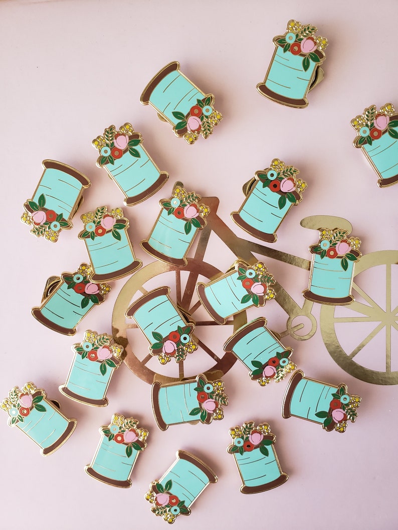 May include: A collection of 18 gold-tone enamel pins featuring a spool of thread with a floral design. The spools are turquoise blue with a gold-tone rim and a variety of colorful flowers.