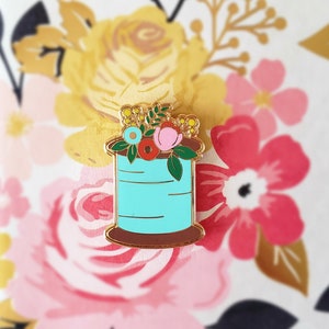 May include: A gold-tone enamel pin featuring a spool of thread with a floral arrangement on top. The spool is turquoise blue with gold accents and a brown base. The flowers are pink, red, yellow, and blue.