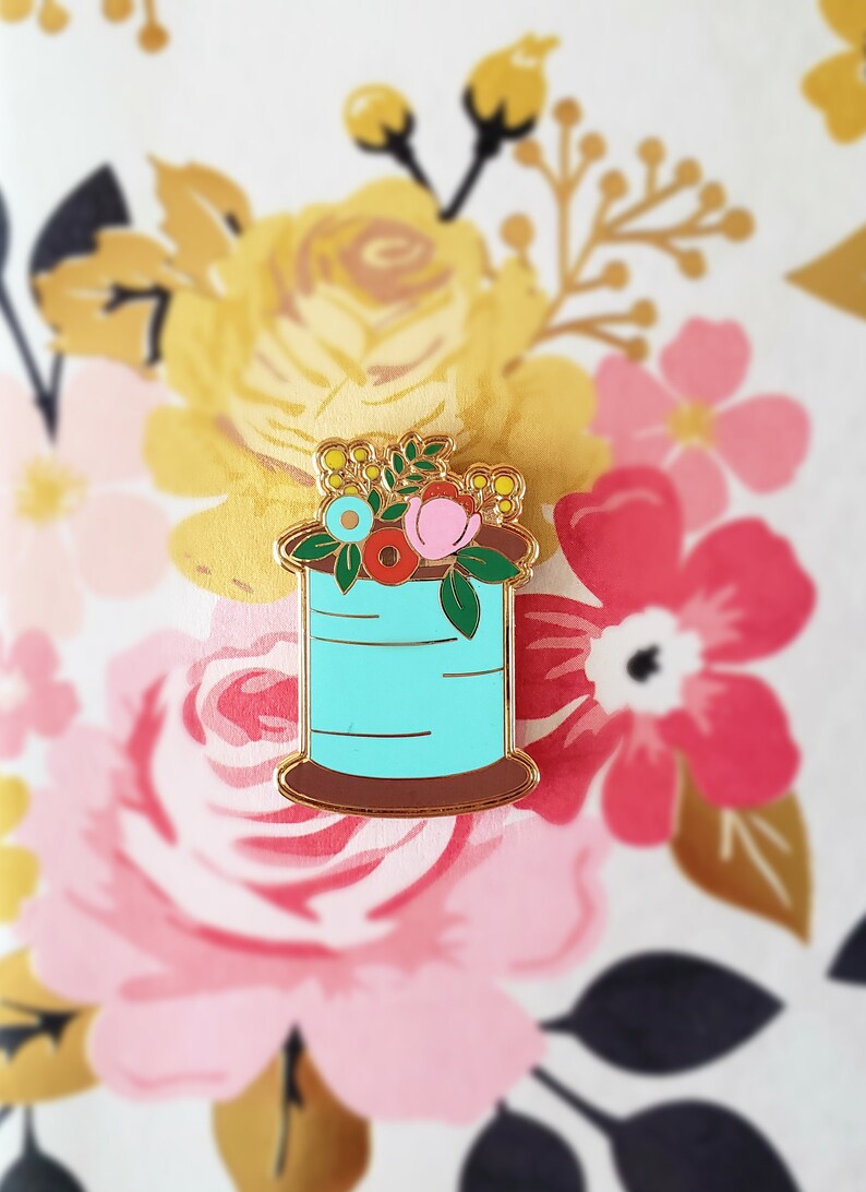 May include: A gold-tone enamel pin featuring a spool of thread with a floral arrangement on top. The spool is turquoise blue with gold accents and a brown base. The flowers are pink, red, yellow, and blue.