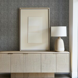 Op de afbeelding: Een licht houten kast met vier deuren en een rechthoekige basis. Een ingelijst abstract kunstwerk en een beige keramische lamp met een witte lampenkap staan erop. De achtergrond is een getextureerde grijze muur.