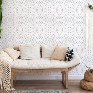 May include: A white sofa with cushions and a throw blanket sits in front of a white wall with a light purple geometric pattern. A macrame wall hanging and a wicker basket with a plant are also visible.