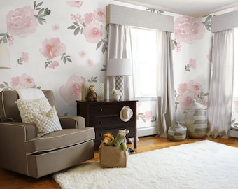 May include: A brown glider rocking chair with a white trim, two pillows, and a white throw blanket. The chair is in a nursery with a white wall featuring a pink floral wallpaper pattern. There is a dark brown dresser with three drawers and a white rug on the floor.