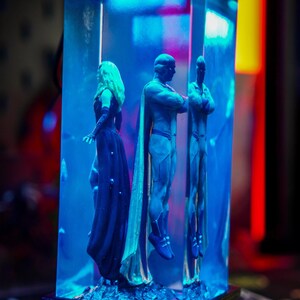 Puede incluir: Una escultura con tres figuras, dos masculinas y una femenina, encerradas en un bloque rectangular transparente. Las figuras son en tonos azules y dorados, con la figura femenina con un vestido largo y oscuro. El fondo est&aacute; iluminado con luz azul.
