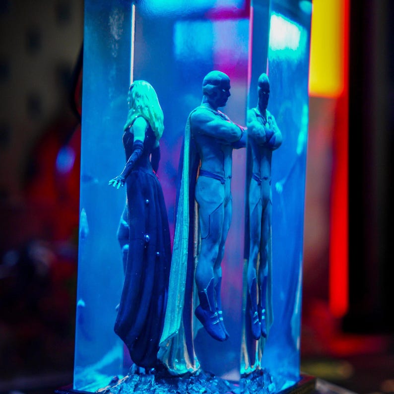 Puede incluir: Una escultura con tres figuras, dos masculinas y una femenina, encerradas en un bloque rectangular transparente. Las figuras son en tonos azules y dorados, con la figura femenina con un vestido largo y oscuro. El fondo est&aacute; iluminado con luz azul.