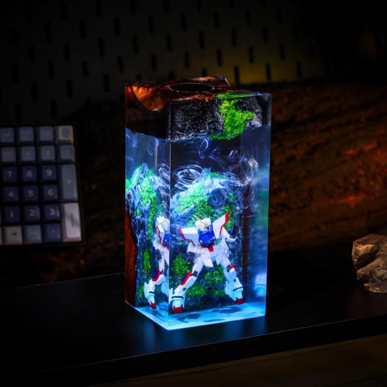 Pode incluir: Uma pe&ccedil;a de arte retangular em resina com uma figura de rob&ocirc; em miniatura. A resina transparente envolve o rob&ocirc;, musgo verde e elementos azuis e brancos em redemoinho. A parte superior &eacute; escura com um buraco e a base brilha em azul.