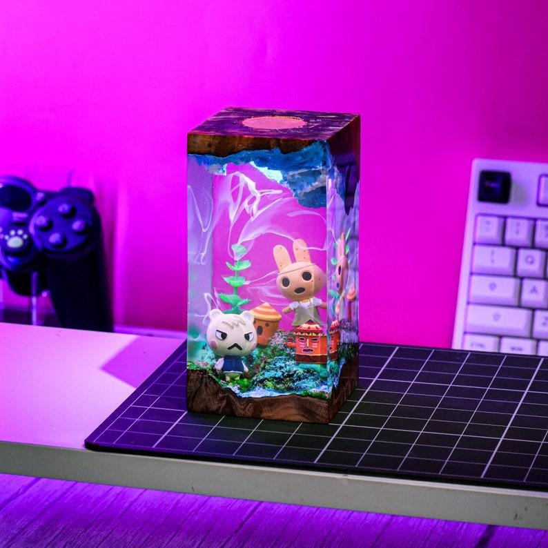 May include: A rectangular resin lamp featuring miniature figurines of characters from a video game. The lamp has a wooden base, blue water effect, and a pink and purple background. The figurines include a rabbit, a cat, and a small house.