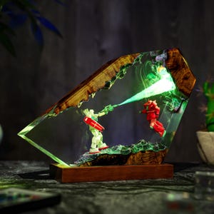 Puede incluir: Una pieza de arte de resina &uacute;nica que presenta dos figuras de robots en una escena de batalla, iluminada por luz verde. La obra de arte est&aacute; encerrada en un bloque de resina transparente con una base y detalles de madera.
