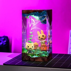 May include: A rectangular resin art piece featuring characters from Animal Crossing. The artwork has a wooden base and a top with blue and white accents. Inside, there are miniature figures of characters, plants, and a small building. The background is a gradient of purple and white.