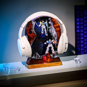 May include: White over-ear headphones resting on a decorative wooden base. The base features a colorful, artistic depiction of a space scene with two robot figures. The headphones and base are on a white surface.