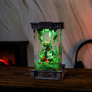 May include: A decorative light-up display featuring video game characters. The display is encased in a clear rectangular structure with a detailed base and top. The interior scene includes figures, green pixelated foliage, and a warm glow.
