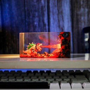 May include: A rectangular resin sculpture featuring a Bowser figure breathing fire. The sculpture has a clear front and sides, with a fiery red and orange interior. The Bowser figure is detailed with green spikes and a black studded collar.