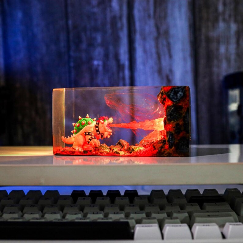 May include: A rectangular resin sculpture featuring a Bowser figure breathing fire. The sculpture has a clear front and sides, with a fiery red and orange interior. The Bowser figure is detailed with green spikes and a black studded collar.