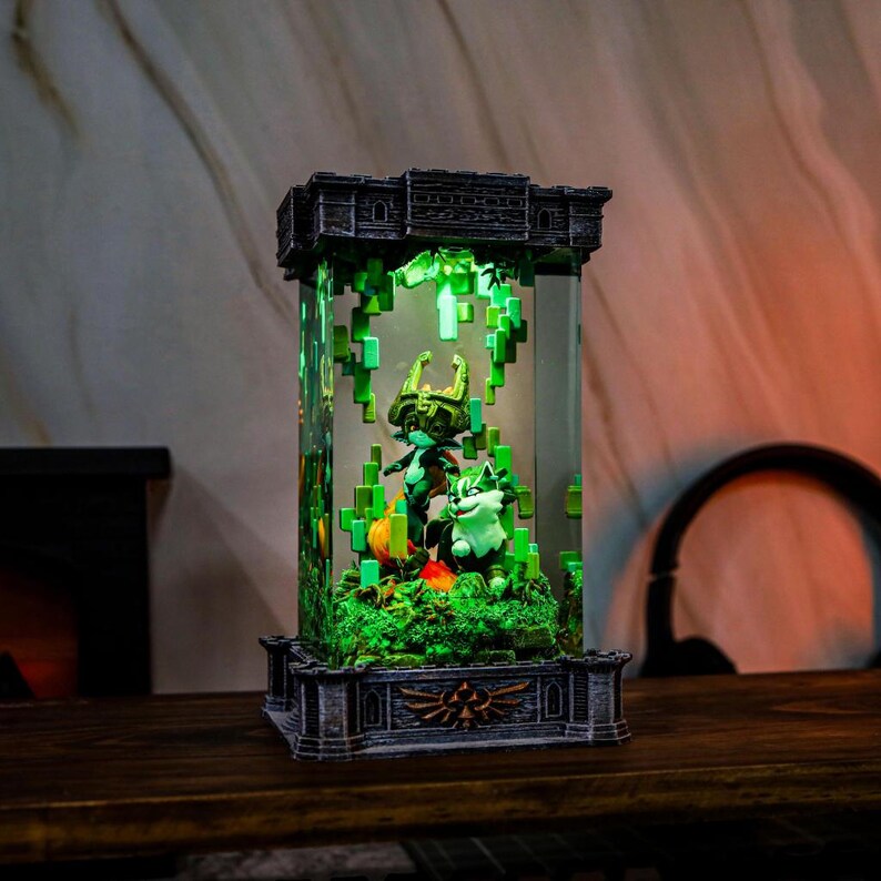 May include: A decorative light-up display with a video game theme. The display has a rectangular glass enclosure with a detailed base and top. Inside, figures of characters are set against a pixelated green background. The base has a gold emblem.
