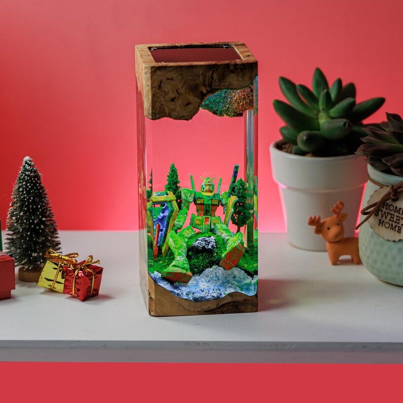 Peut inclure: Une pi&egrave;ce d'art en r&eacute;sine rectangulaire avec un dessus et une base en bois, repr&eacute;sentant un robot vert dans un paysage miniature. Le robot est entour&eacute; de petits arbres et d'une base bleue, le tout enferm&eacute; dans de la r&eacute;sine transparente.
