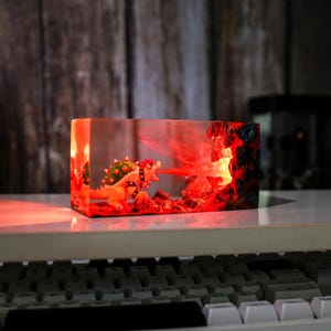 May include: A rectangular resin sculpture depicting Bowser breathing fire. The sculpture is lit with red light, creating a fiery effect. The background has a wooden texture.