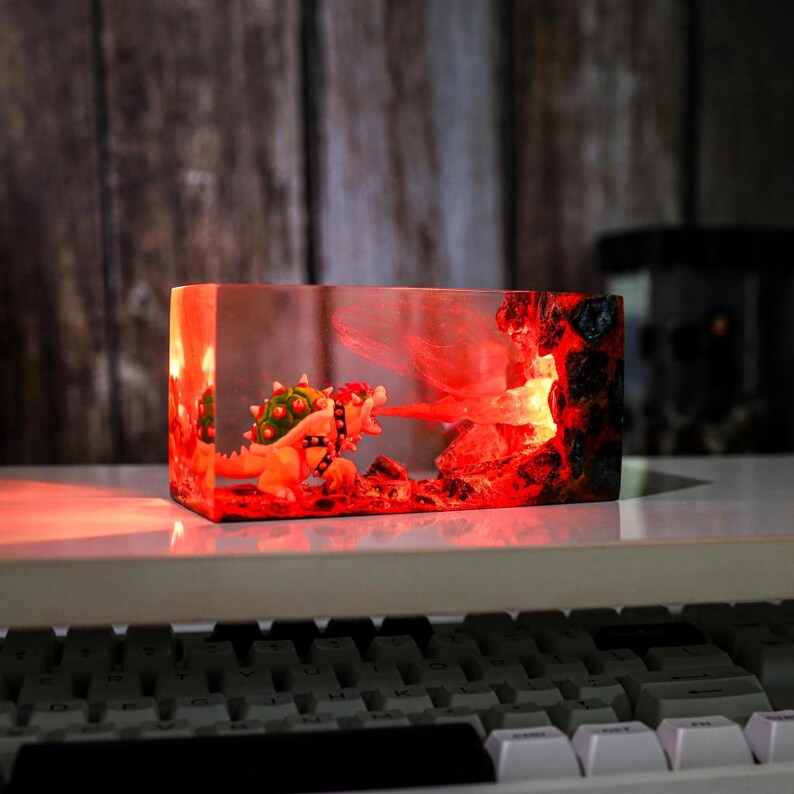 May include: A rectangular resin sculpture depicting Bowser breathing fire. The sculpture is lit with red light, creating a fiery effect. The background has a wooden texture.