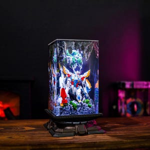 May include: A rectangular LED lamp featuring a detailed model of a robot warrior. The lamp has a black base and a clear, illuminated body with a colorful design. The robot is white, red, blue, and yellow. The background shows a cave-like structure.