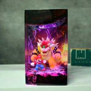 Op de afbeelding: Een rechthoekig kunstwerk van hars met de personages Mario en Bowser uit de Super Mario-videogame serie. Bowser is een groot, oranje schildpadachtig wezen met stekels, en Mario draagt zijn klassieke rode en blauwe outfit. De achtergrond heeft een paarse en oranje gloed.