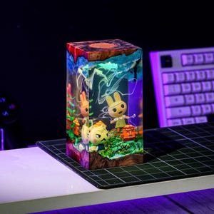 May include: A rectangular resin block featuring miniature Animal Crossing characters in a diorama. The top has a wood grain appearance, while the sides are clear, showcasing the characters and colorful details. The scene is illuminated with a soft glow.