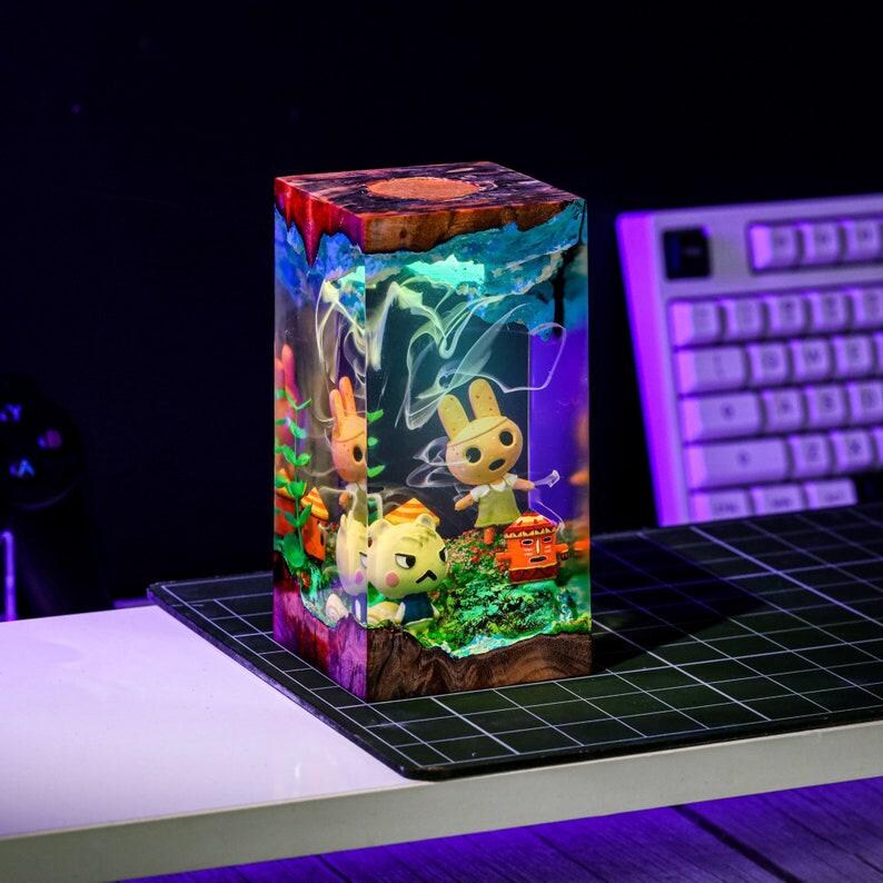 May include: A rectangular resin block featuring miniature Animal Crossing characters in a diorama. The top has a wood grain appearance, while the sides are clear, showcasing the characters and colorful details. The scene is illuminated with a soft glow.
