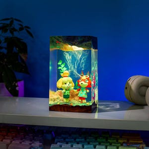 May include: A rectangular resin lamp featuring two cartoon characters in a simulated underwater scene. The lamp has a light source inside, illuminating the figures and the surrounding environment. The characters are a yellow dog and a red fox.