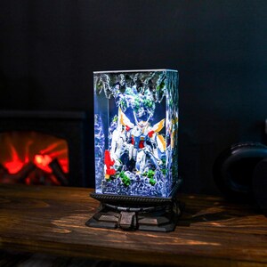 May include: A rectangular, illuminated display featuring a detailed robot model with white, red, yellow, and blue accents. The model is set within a clear, textured enclosure, mounted on a decorative, dark-colored base.