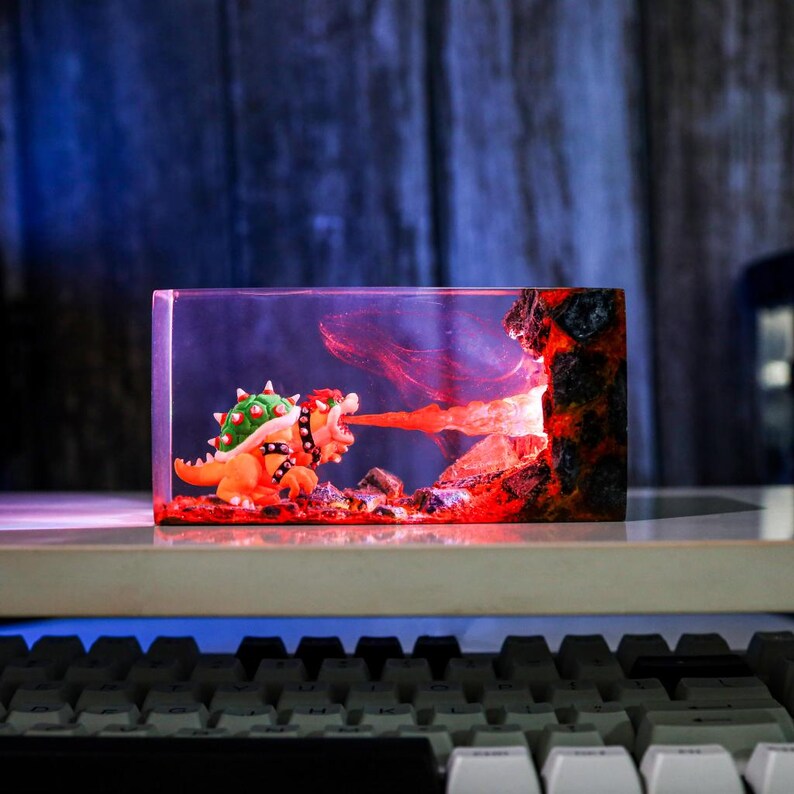 May include: A resin diorama featuring a Bowser figure breathing fire. The scene is set within a clear rectangular block, with a fiery red and orange colour scheme. The figure is orange and green, with a black collar. The background has a volcanic rock design.