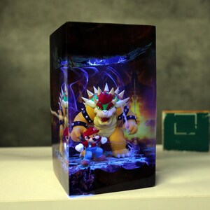 Op de afbeelding: Een rechthoekige, oplichtende display met de personages Bowser en Mario uit de Super Mario-videogame serie. Bowser is een groot, geel, schildpadachtig wezen met een rode hanenkam. Mario is een kleine figuur met een rode hoed en blauwe overall.