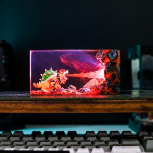 May include: A resin diorama featuring a Bowser figure breathing fire. The scene is set inside a rectangular block with a volcanic rock formation on the right. The figure is orange and green, with a black studded collar. The background is a dark purple.