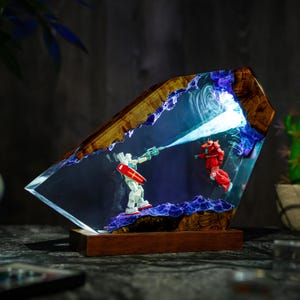 Puede incluir: Una pieza de arte de resina &uacute;nica que presenta dos figuras de robots en una escena de batalla. La obra de arte est&aacute; encerrada en una forma geom&eacute;trica transparente con un marco y una base de madera. El interior tiene detalles en azul y blanco.