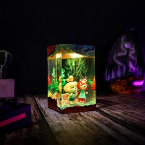 May include: A rectangular resin lamp featuring two cartoon characters in a miniature diorama. The lamp has a wooden base and a clear resin body, with green and red plant life. The characters are a yellow dog and a red fox, both wearing outfits.